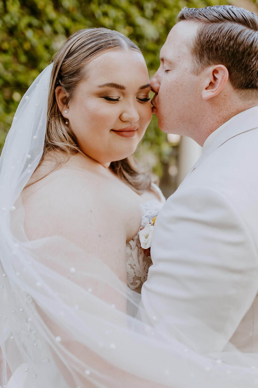 groom kisses brides temple while her veil flows in the wind