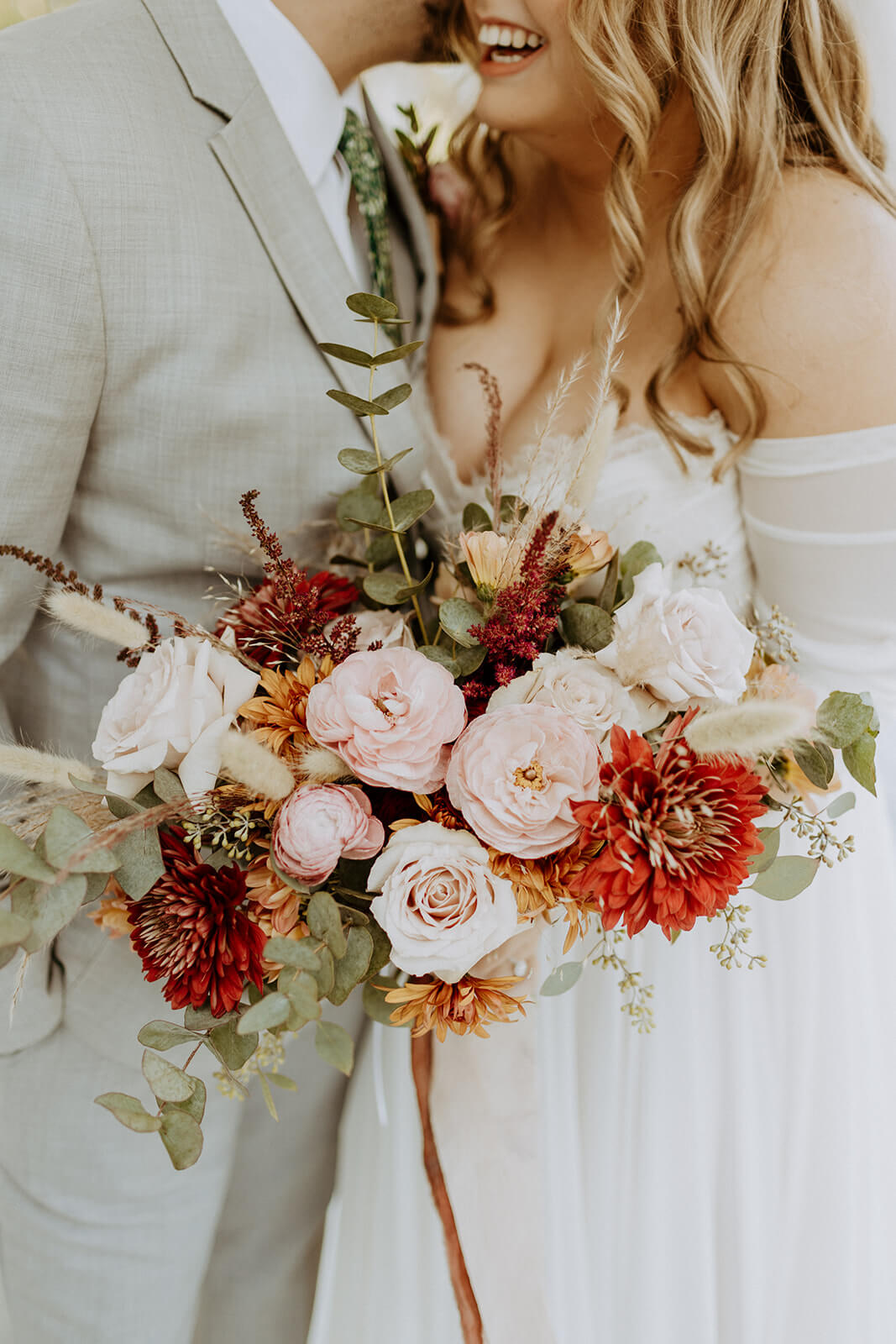 close up image of brides fall colored floral arrangement while she laughs with husband