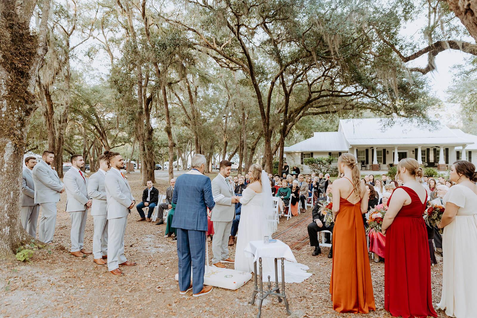 bride and groom stand at alter surrounded by wedding guests and wedding party under large moss covered trees